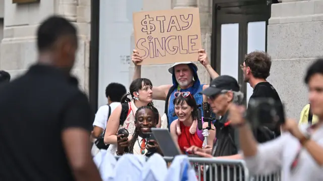 A group of people stands behind a metal fence, with a man holding up a sign reading "stay single" on it