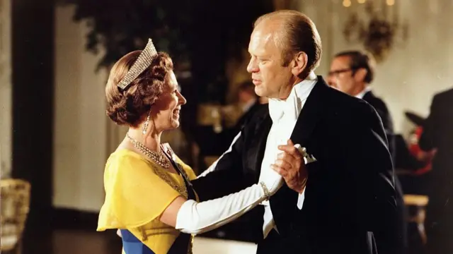 Queen Elizabeth II (L) in a yellow dress and blue sash across her chest, the Queen Alexandra Kokoshnik Tiara on her head, dances with US President Gerard Ford (R) in a black tuxedo at the White House