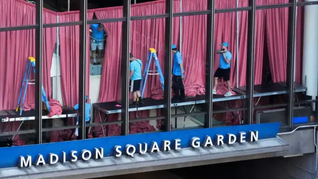 People in blue t-shirts stand on ladders putting up red curtains behind some tall windows