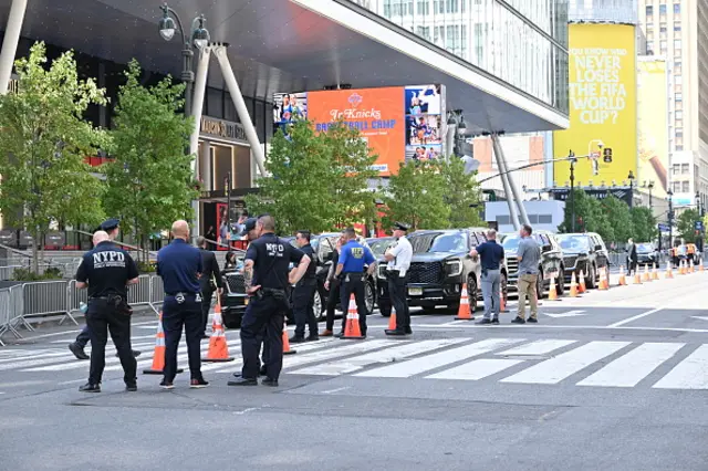 Black cars with blacked out windows line up next to several police officers. There are barriers on one side of the road and cones on the other