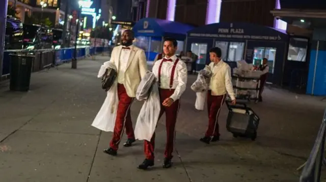 Three men in cream jackets and red trousers walk down the street, one wheels a cart behind him.