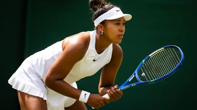 Naomi Osaka, wearing a white dress and visor with her dark hair in a bun, faces right and stands bent over her blue racquet waiting to receive a serve