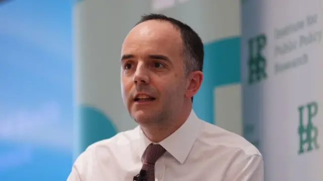 Mid-shot of Health Secretary James Murray sitting down at an event. He's in a white shirt and brown patterned tie. In the blurred background is a white sign with the words Institute for Public Policy Research in green
