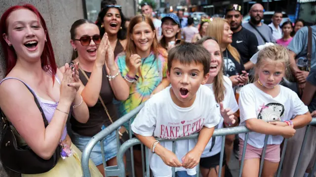 A group of women and children are seen stood behind a metal barrier, with most smiling and clapping