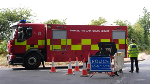 A fire engine travels along a road that is cordoned off by a single female police officer who is wearing high-vis and black trousers.There is a blue sign that reads POLICE ROAD CLOSED