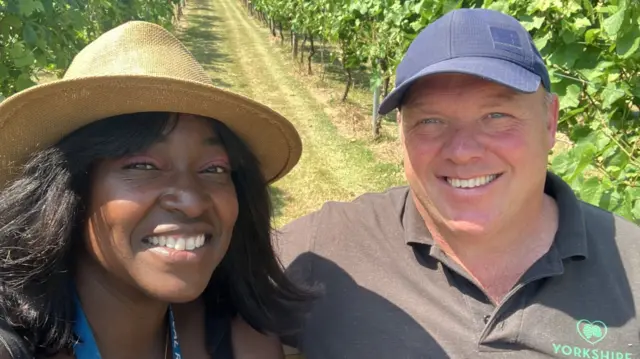 BBC reporter Joanita Musisi smiles in a hat next to Tim Spakouskas, who works on the vineyard