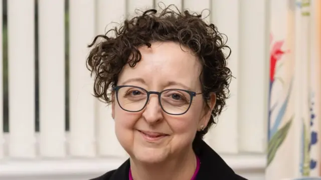A head and shoulders image of a female bishop. She is sitting on a chair with blinds and curtains in the background. She is wearing glasses and a black blazer over a pink top with her clerical collar on. She is looking directly into the camera.