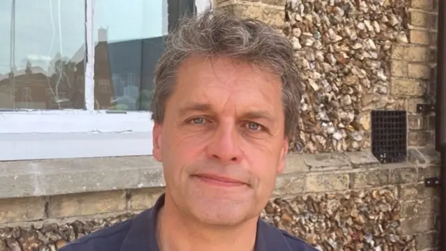 A man looks at the camera slightly smiling. He has dark hair with grey in it. He is wearing a blue polo T-shirt.