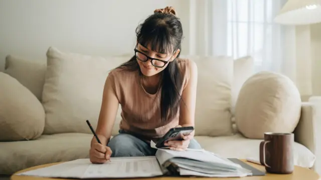 A woman sits at her living room with smartphone and paperwork doing her monthly budget