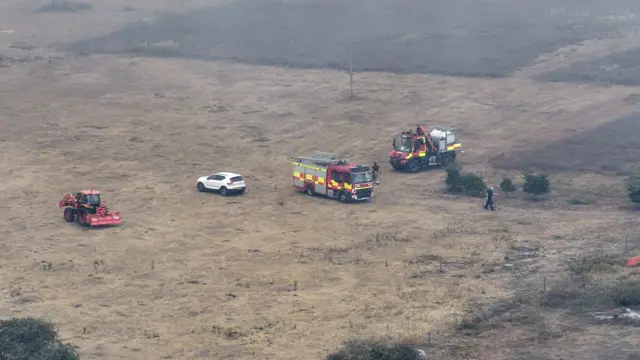 A drone image of heathland that has been burnt in a fire. Several fire engine vehicles are parked in a field along with a tractor and white vehicle. Some firefighters are walking around the area.