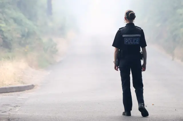 The back of a woman in black police uniform. White smoke drifts across a single lane, with bushes either side on the steep banks.