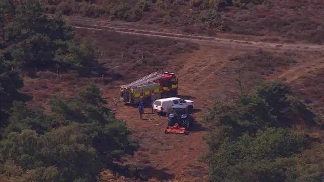 A bird's eye or drone view of a red and yellow fire engine, and a white car, parked on a sandy heathland, with some green bushes around them.