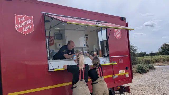 A red Salvation Army Emergency Response Vehicle. A man is leaning over it open hatch passing drinks to two firefighters. They are leaning up to receive them.