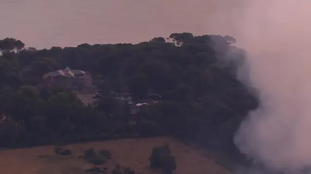 A bird's eye view of green trees and a large building, but white smoke billows in the foreground. The sea is visible.