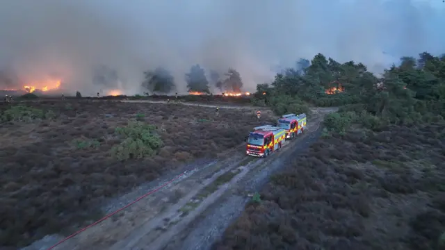 Two red and yellow fire engines are on a unmade track of byway, with bushes either side, and there are red flames in the background.