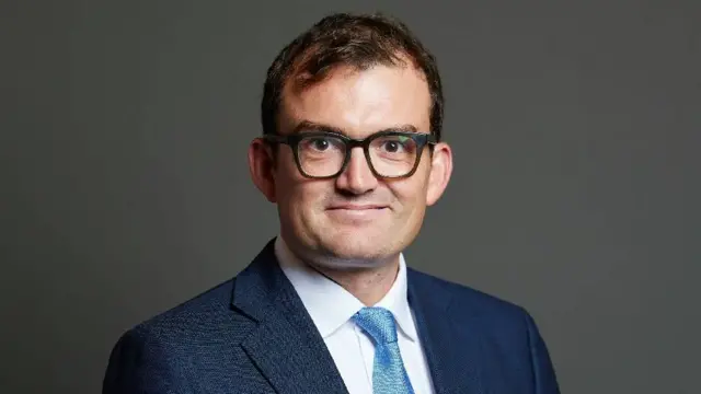 Patrick Spencer wears a navy suit and stands in front of a plain background. He is smiling directly at the camera and is wearing large black-rimmed glasses.