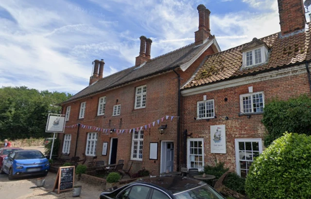 The red brick exterior of a village pub where bunting has been placed across its front. It is two-storey, and there are cars parked outside. There is blue sky and white clouds above.