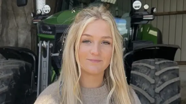 Harriet Cowan, a young woman, smiles at the camera. She has long blonde hair and is wearing a light brown jumper. Behind her is a tractor