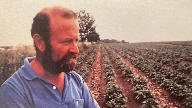 Photo of Mike Potter standing on a farm wearing a blue T-shirt. Side profile with rows of crops in the background, it looks like an old and slightly faded photo.