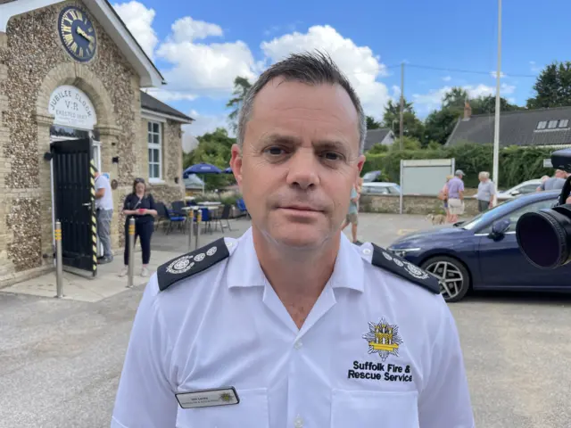 Jon Lacey wears a white shirt with black epaulettes on the shoulders and a yellow crest on the chest, with the words printed SUFFOLK FIRE & RESCUE SERVICE. He is outside in a car park.