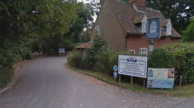 An entrance and road into a holiday park. There is a lone house on the side with various entry signs.