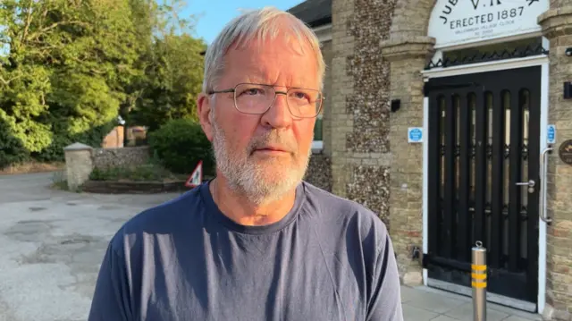A man with a white beard, white hair and glasses in a grey t-shirt outside a village hall.