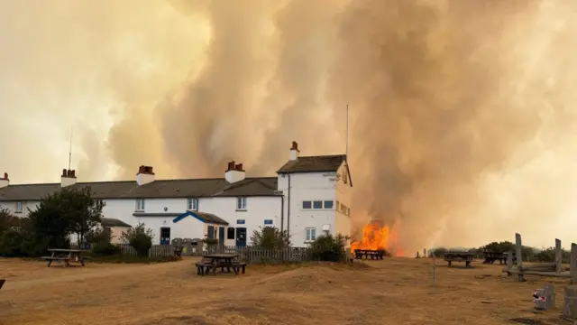 Fire burns near to cottages. Thick smoke can be seen billowing into the air.