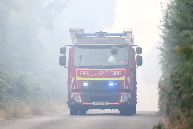 A fire engine travels along a road with its emergency lights going off. Smoke fills the air. Bushes and trees line the road.