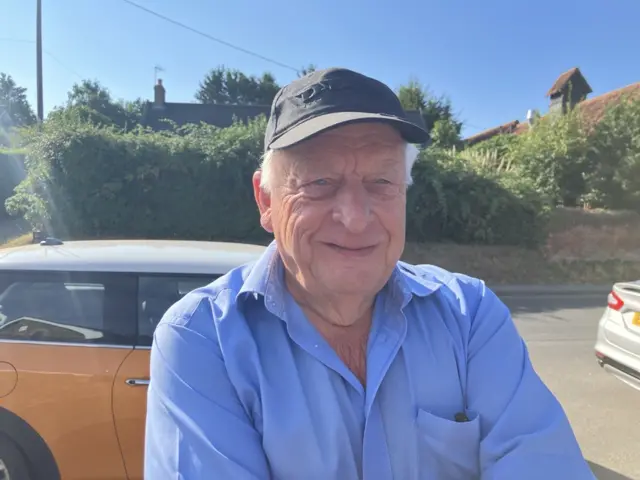 Colin Fisher stands in front of a car, he is wearing a dark baseball cap and light blue shirt.