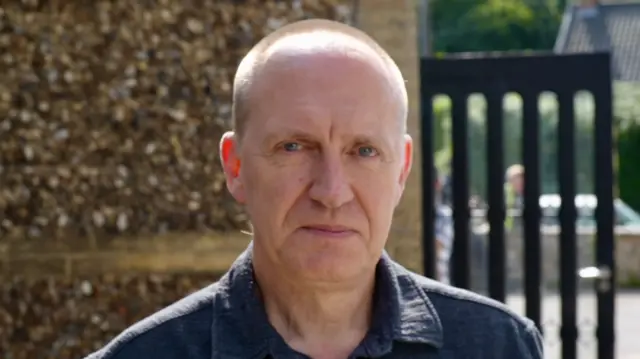 A man standing outside looks at the camera. He is bald and wearing a dark blue shirt.