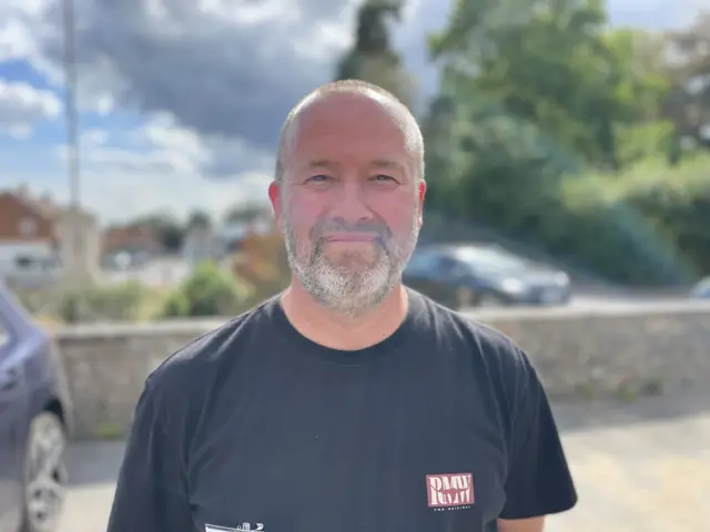 Barry Banks has very short hair and has a beard. He wears a navy blue T-shirt. He is outside with stone brick wall behind him.
