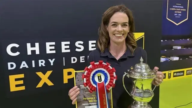 Lowri Wright holds a trophy in one hand and a rosette and certificate in the other. She has long brown hair and is standing in front of signage saying Cheese Dairy Expo