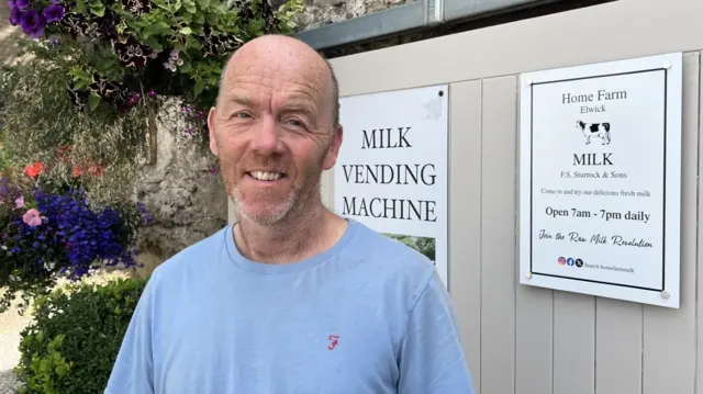 A bald man stands in front of a sliding wooden door of a grey brick building. He is smiling at the camera and wearing a blue t-shirt. Behind him is a white sign which says "milk vending machine", to the right of this is another sign which says "Home Farm Elwick, Milk".