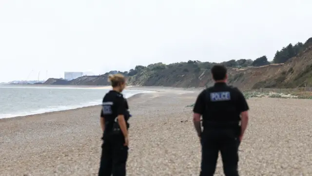 Two uniformed officers stand on a beach with their backs to the camera. On their right is a coastal cliff and in the distance Sizewell nuclear power station can be seen.