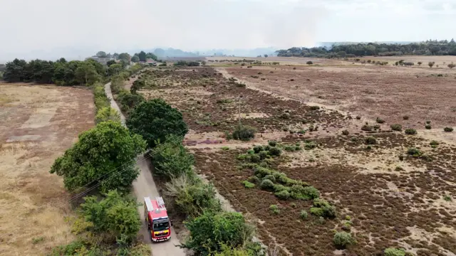 A red and yellow branded fire engine drives towards the camera on a narrow road. Either side of the road is brown dry earth. In the background is white billowing smoke.