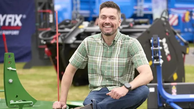 A man sit smiling at the camera. He has short brown hair, a short beard and is wearing a green checked shirt and blue jeans. Behind him is farming equipment