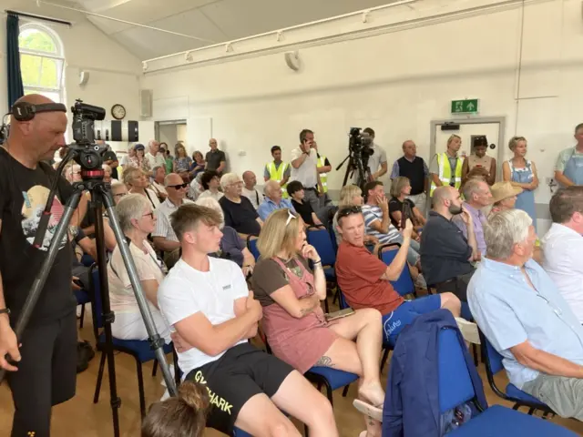 Dozens of people sit in seats in a town hall. A camera operator wear a black T-shirt and pointing a camera is to the left.