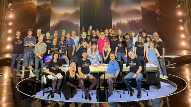 A large group of people stand in tiers around the Britian's Got Talent judges table. The stage is empty and dark but there are bright spotlights on the sides