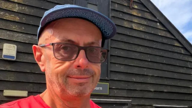 A man smiles at the camera. He wears a blue hat, sunglasses and red T-shirt.
