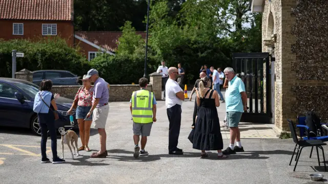 Members of the public talk while standing outside a village hall on a sunny day. There chairs are dotted about the place and some officials wearing hi-vis tops.