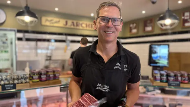 Jamie Archer holds a cut of beef and a bottle of beer inside Archers Butchers in Norwich