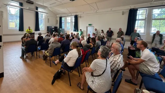 People gathered in a hall with a man in a high vis jacket giving information from a podium.