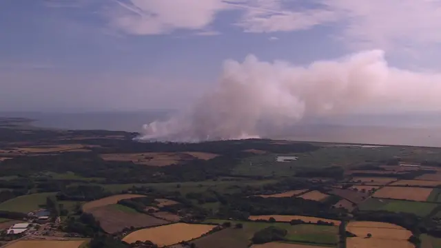 Large plumes of white smoke are billowing from an area of fields beside the sea. The photo is taken from above, perhaps from a helicopter.