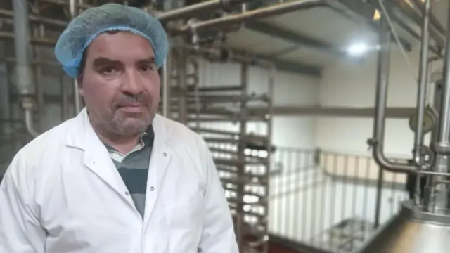 Mathieu Varley, a man dressed in a white coat and blue hairnet, stands among metal milk churns and pipes in a dairy. He has brown hair and grey stubble.