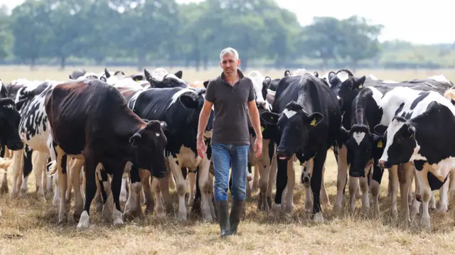 A man with grey hair, dark T-shirt, jeans and wellies looks at the camera, standing in front of a herd of black and white cows in a field, trees blurred in the background