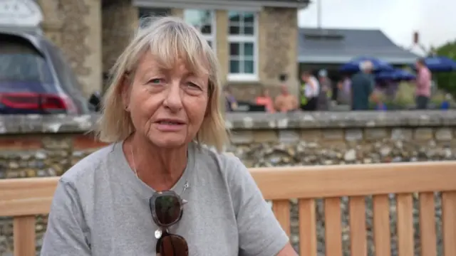 Jude sat on a wooden bench outside a village hall. She wears a grey T-shirt, has blonde hair and has sunglasses hanging from her collar.