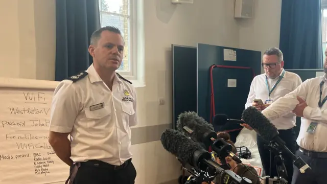 A fire chief wearing a white shirt and black trousers stands in front of various microphones. He has his hands behind his back. He has short dark hair. There are journalists standing in front of him listening.