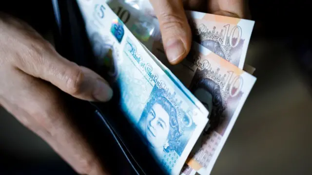 Hands put pound sterling notes into a wallet. Queen Elizabeth II's face is visible on the notes.