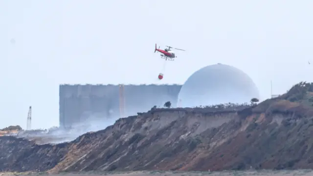 A helicopter carrying a suspended large water bucket above a coastal area toward a fire. A large dome shaped building part of a nuclear power station can be seen in the distance behind the helicopter.