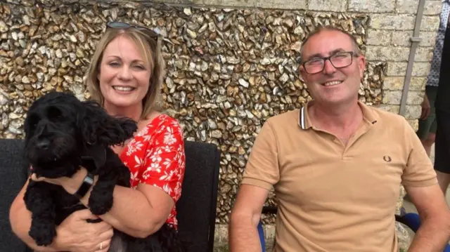 A man and a woman sit next to each other on chairs outside a building. They are smiling at the camera. The woman has short blonde hair and has a black dog on her lap. She is also wearing a red dress with white flowers on it. The man has short hair and wears a light brown polo top. He is holding a cup of coffee.